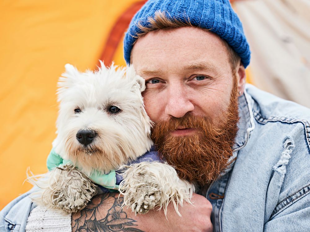 Man with a knit hat and beard holding a white terrier up to his face in an endearing snuggle